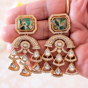 May include: Gold-toned chandelier earrings with emerald-green and clear gemstone accents. The earrings feature rectangular emerald-green stones at the top, surrounded by smaller clear stones. Below, a curved design is adorned with more clear stones, leading to dangling teardrop and triangle-shaped clear stones.