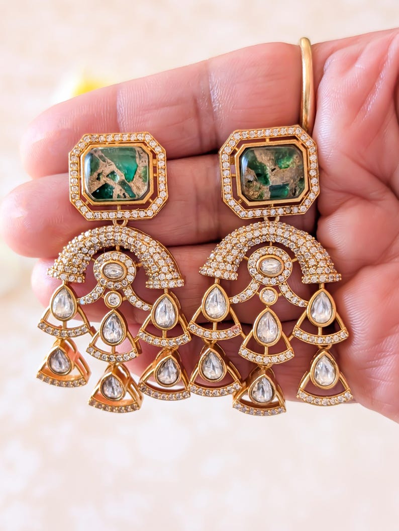 May include: Gold-toned chandelier earrings with emerald-green and clear gemstone accents. The earrings feature rectangular emerald-green stones at the top, surrounded by smaller clear stones. Below, a curved design is adorned with more clear stones, leading to dangling teardrop and triangle-shaped clear stones.
