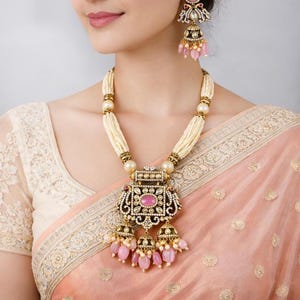 May include: A jewelry set consisting of a necklace and matching earrings. The necklace features multiple strands of cream-colored beads, a large rectangular pendant with a central pink stone, and three dangling bell-shaped charms with pink beads.