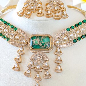 May include: A gold-toned jewelry set featuring a necklace and matching earrings. The necklace has a large, rectangular green stone pendant and smaller green stones along the band. The earrings and necklace have a cascading design with clear, teardrop-shaped stones.