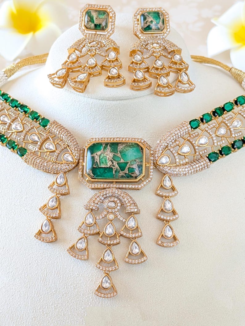 May include: A gold-toned jewelry set featuring a necklace and matching earrings. The necklace has a large, rectangular green stone pendant and smaller green stones along the band. The earrings and necklace have a cascading design with clear, teardrop-shaped stones.