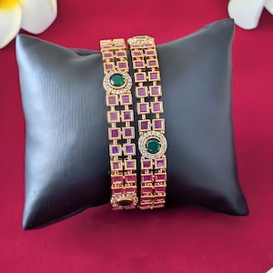 May include: Two gold bangles with a square design and purple and green stones. The bangles are on a black pillow.