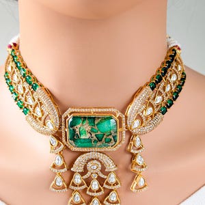 May include: A gold-toned necklace with a large rectangular green stone center. The necklace features intricate detailing with clear and green gemstones. The design includes teardrop and arch-shaped elements, creating a luxurious and ornate appearance.
