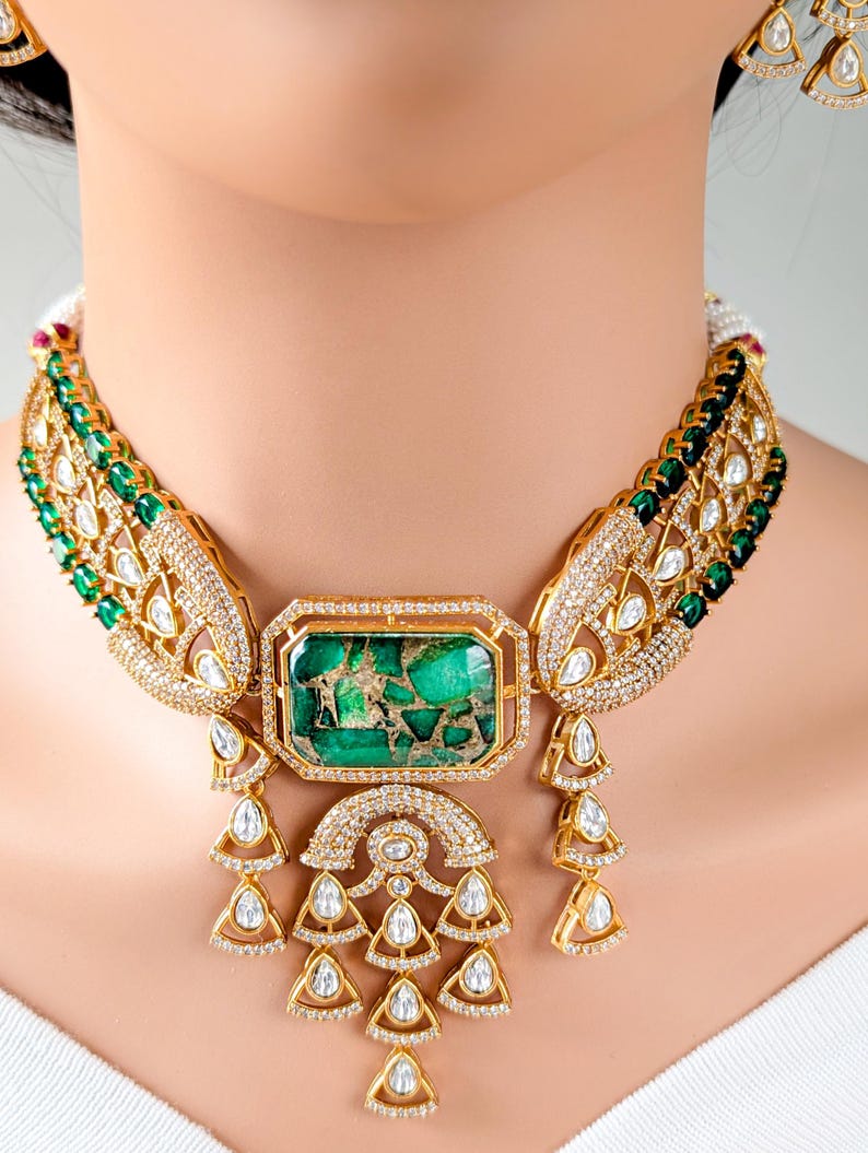 May include: A gold-toned necklace with a large rectangular green stone center. The necklace features intricate detailing with clear and green gemstones. The design includes teardrop and arch-shaped elements, creating a luxurious and ornate appearance.