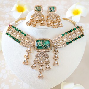 May include: A gold-toned necklace and earring set featuring emerald green and clear gemstones. The necklace has a central rectangular green stone and dangling teardrop accents. The earrings mirror the necklace design, with square green stones and cascading details. The jewelry is displayed on a white stand.