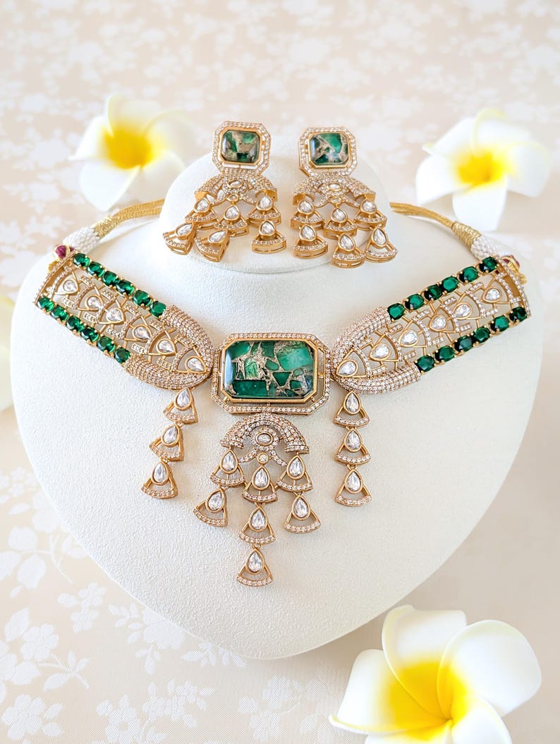 May include: A gold-toned necklace and earring set featuring emerald green and clear gemstones. The necklace has a central rectangular green stone and dangling teardrop accents. The earrings mirror the necklace design, with square green stones and cascading details. The jewelry is displayed on a white stand.