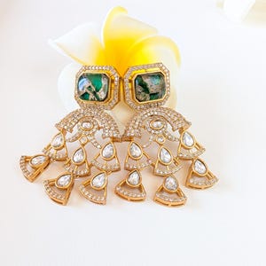 May include: A pair of gold-toned earrings with emerald-green square center stones. The earrings feature a cascading design of clear, round and teardrop-shaped crystals. The earrings are set against a white background with a yellow and white flower.