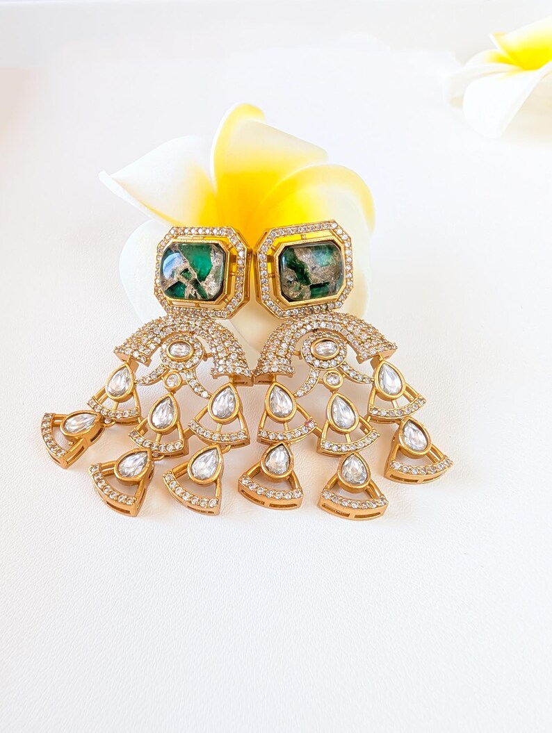 May include: A pair of gold-toned earrings with emerald-green square center stones. The earrings feature a cascading design of clear, round and teardrop-shaped crystals. The earrings are set against a white background with a yellow and white flower.