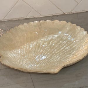 May include: A decorative dish shaped like a large scallop shell with a glossy, off-white, pearlescent finish. The surface features radiating ridges and a scalloped edge, mimicking the natural texture and form of a seashell in a smooth, hard material.