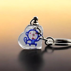 May include: A clear heart-shaped keychain with a blue monkey design inside. The monkey is holding a star and has pink cheeks. The keychain is attached to a silver metal ring.