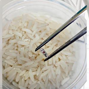 May include: A clear glass bowl filled with white rice. A pair of tweezers holds a small white strip of paper with the words "I ❤️ You" printed on it. The image is well-lit, with a focus on the rice and the message.