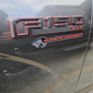 Puede incluir: Una camioneta Ford F-150 XLT negra con un emblema rojo y negro que dice "F-150 XLT" y una pegatina negra y gris que dice "SUPERCHARGED" con un gráfico de lobo.