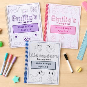 May include: Three spiral-bound tracing books with white covers. Two books are titled Emilia's Tracing Book with purple and pink accents, featuring Halloween and flower graphics. One book is titled Alexander's Tracing Book with blue accents and dinosaur graphics.