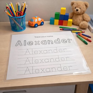 May include: A worksheet with the name "Alexander" printed in dotted letters for tracing, with the text "Trace your name" above. The worksheet is on a wooden desk with colorful pencils, crayons, and toys.