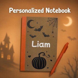 May include: A personalized notebook with the name "Liam" printed on the cover. The cover features a Halloween theme with a pumpkin, bats, a spider web, and a crescent moon. An orange pen rests beside the notebook.