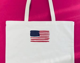 May include: White tote bag with a printed American flag design. The flag features a blue rectangle with white stars and red and white stripes. The bag has white handles and is made of a canvas-like material.