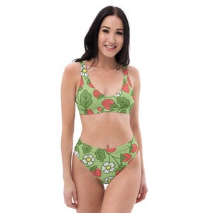 Puede incluir: Un traje de baño de dos piezas con un fondo verde claro, adornado con un estampado de fresas rojas, flores blancas y hojas verdes. La parte superior es de cuello redondo y la parte inferior es de cintura alta.