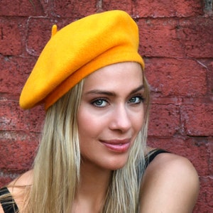 May include: A woman wearing a bright yellow beret. The beret is made of felt and has a classic style. The woman has long blonde hair and is wearing a black tank top.