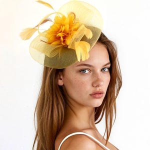 May include: A yellow fascinator hat featuring a circular mesh base and a central floral accent with delicate feathers. The headpiece is constructed from sheer, layered netting material in a bright, sunny yellow hue, creating a structured yet airy appearance.