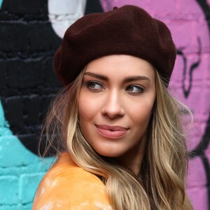 May include: A woman with long blonde hair wearing a brown beret and a yellow shirt. She is standing in front of a colorful brick wall.