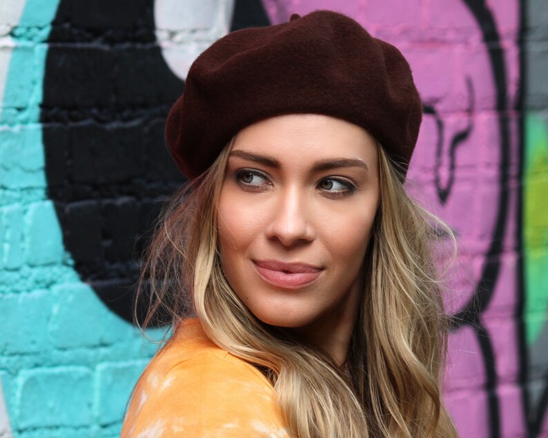 May include: A woman with long blonde hair wearing a brown beret and a yellow shirt. She is standing in front of a colorful brick wall.