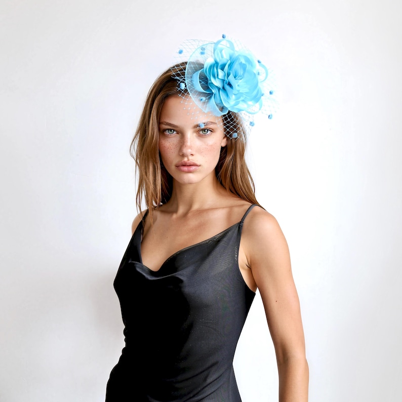 May include: A bright turquoise fascinator headpiece featuring a large, layered fabric flower and a delicate mesh netting veil. The accessory is adorned with small, matching turquoise beads scattered throughout the netting, creating a textured look.