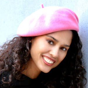 May include: A woman with curly brown hair is wearing a pink beret. She is smiling and looking at the camera.