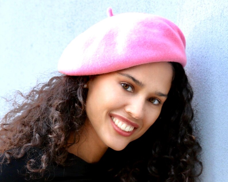 May include: A woman with curly brown hair is wearing a pink beret. She is smiling and looking at the camera.