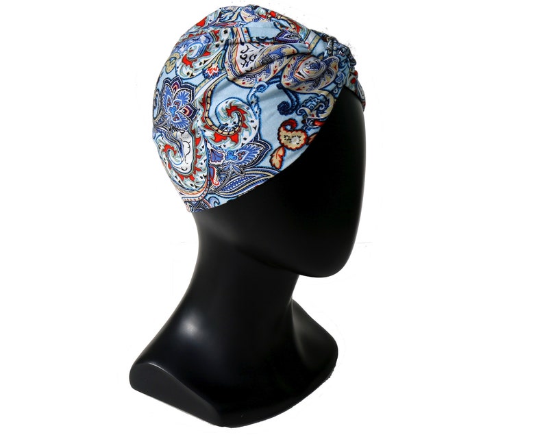May include: A light blue turban with a paisley pattern in red, white, and blue. The turban is displayed on a black mannequin head, showcasing the head covering's design and fit. The fabric appears soft and lightweight.