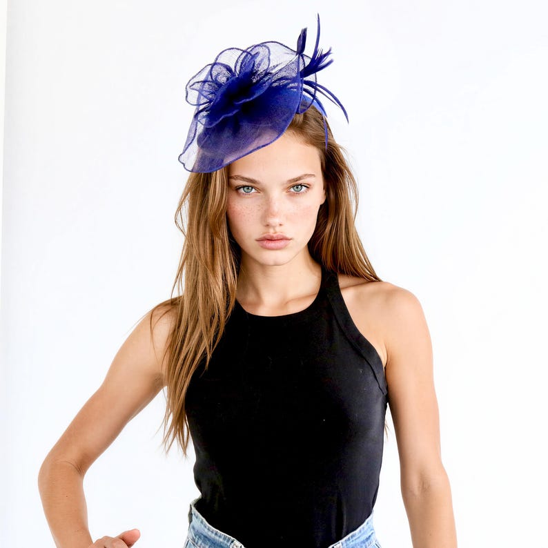 May include: A dark blue fascinator with a floral design and feather accents. The headpiece is made of sheer fabric and is worn on the head. The background is white.