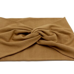 May include: A tan-colored, knotted headband made of soft fabric. The headband is a solid color and has a twisted knot detail at the center. The fabric appears to be a knit or jersey material, suitable for casual wear.