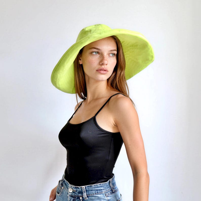 May include: A wide-brimmed, lime green sun hat. The hat has a large, floppy brim and a rounded crown. The hat is made of a soft, textured fabric. The model is wearing a black tank top and blue jeans.