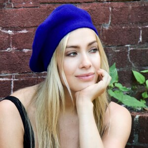 May include: A woman with long blonde hair is wearing a blue beret. She is looking off to the side and has her chin resting on her hand. She is wearing a black tank top and is standing in front of a brick wall.