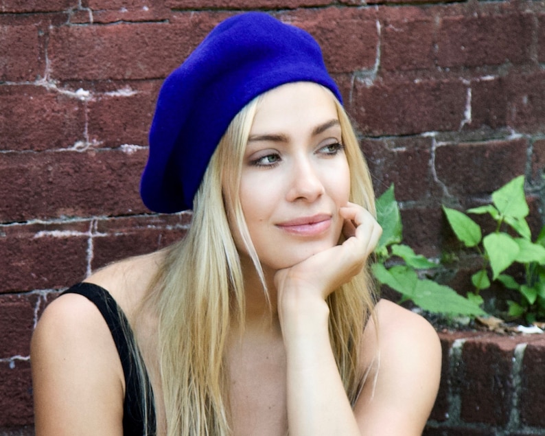 May include: A woman with long blonde hair is wearing a blue beret. She is looking off to the side and has her chin resting on her hand. She is wearing a black tank top and is standing in front of a brick wall.