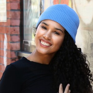 May include: A light blue ribbed knit beanie. The beanie is worn by a person with dark curly hair and a black top. The background includes a brick wall and a painted wall.