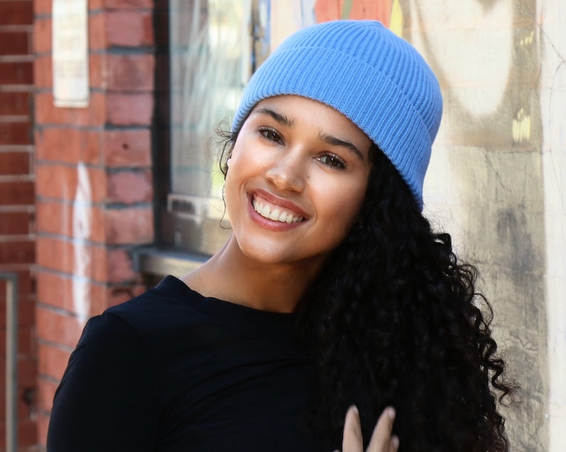 May include: A light blue ribbed knit beanie. The beanie is worn by a person with dark curly hair and a black top. The background includes a brick wall and a painted wall.