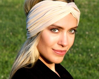 May include: A woman with blonde hair is wearing a light beige turban headband. She is looking at the camera and smiling.
