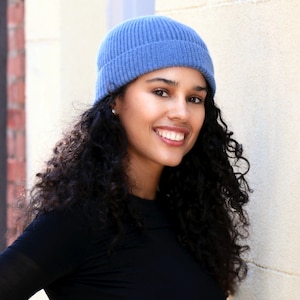 May include: A woman wearing a blue ribbed knit beanie and a black long-sleeve shirt. The beanie is a solid light blue color. The woman has curly dark hair and is smiling.