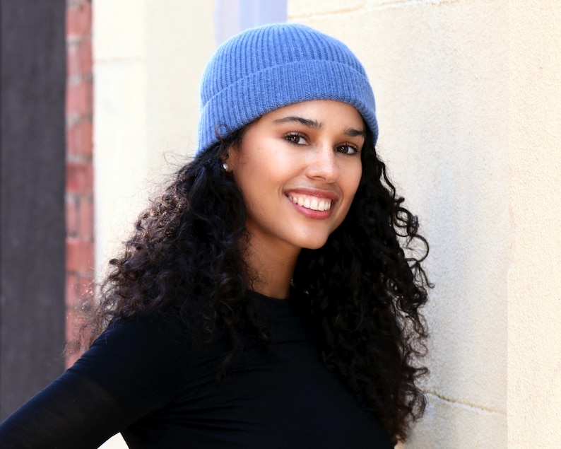 May include: A woman wearing a blue ribbed knit beanie and a black long-sleeve shirt. The beanie is a solid light blue color. The woman has curly dark hair and is smiling.