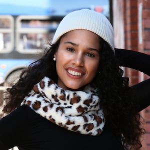May include: A woman wearing a cream-colored knit beanie and a black long-sleeved shirt. She is also wearing a faux fur infinity scarf with a brown and white animal print. The background includes a brick wall and a blurred bus.