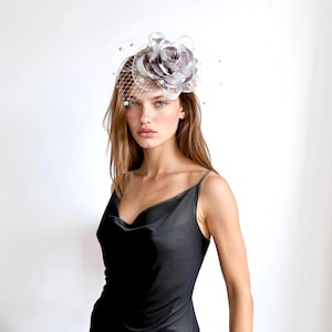 May include: A silver-grey fascinator headpiece featuring a large, layered fabric flower and a delicate netting veil. The accessory is adorned with small, scattered beads and is positioned on the side of the head, paired with a black slip dress.