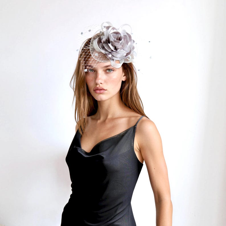 May include: A silver-grey fascinator headpiece featuring a large, layered fabric flower and a delicate netting veil. The accessory is adorned with small, scattered beads and is positioned on the side of the head, paired with a black slip dress.