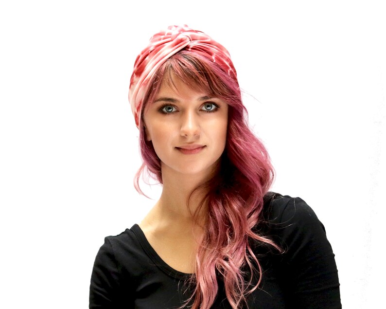 May include: A pink and white turban-style head covering. The head wrap is made of a soft, silky material. The woman in the image is wearing a black top and has pink hair.