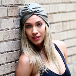 May include: A gray turban-style hat with a knotted design. The hat is made of a soft fabric and is worn by a person with blonde hair. The person is leaning against a brick wall. The hat is a stylish accessory.