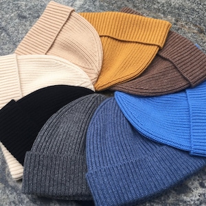 May include: A collection of ribbed knit beanies in various colors, including beige, cream, mustard yellow, brown, black, gray, denim blue, and bright blue. The hats are folded at the brim and arranged in a circular pattern.
