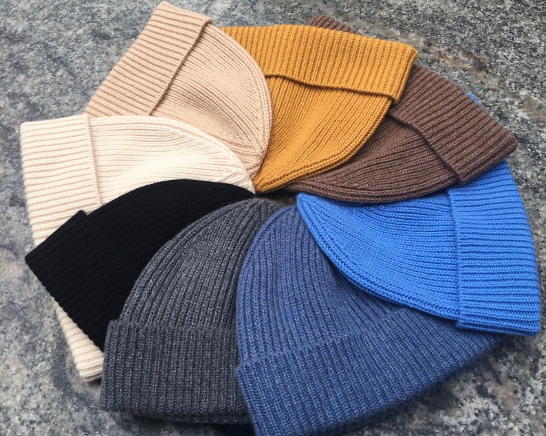 May include: A collection of ribbed knit beanies in various colors, including beige, cream, mustard yellow, brown, black, gray, denim blue, and bright blue. The hats are folded at the brim and arranged in a circular pattern.