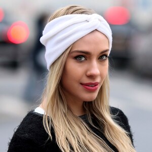 May include: A woman with long blonde hair is wearing a white turban headband. The headband is made of a soft, stretchy fabric and has a knotted design.