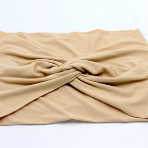 May include: A tan-colored, knotted headband made of soft fabric. The headband is folded and twisted at the center, creating a stylish and comfortable accessory. This hair accessory is suitable for various hairstyles.