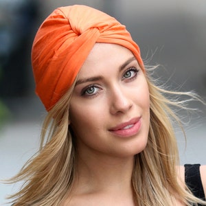 May include: An orange turban-style head covering. The fabric is gathered and twisted at the top, creating a stylish and textured look. The head covering is made of a soft material and is designed to be worn on the head.