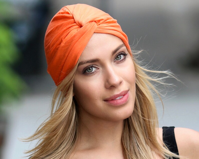May include: An orange turban-style head covering. The fabric is gathered and twisted at the top, creating a stylish and textured look. The head covering is made of a soft material and is designed to be worn on the head.